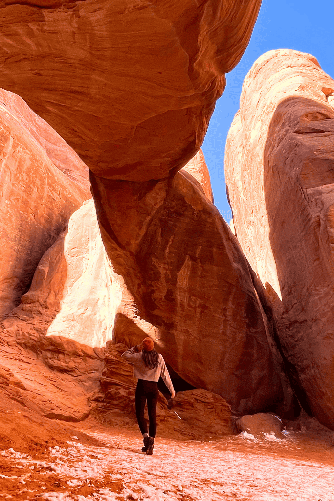 Kate walks beneath a narrow sandstone arch surrounded by towering red rock walls and patches of snow in a shaded canyon.