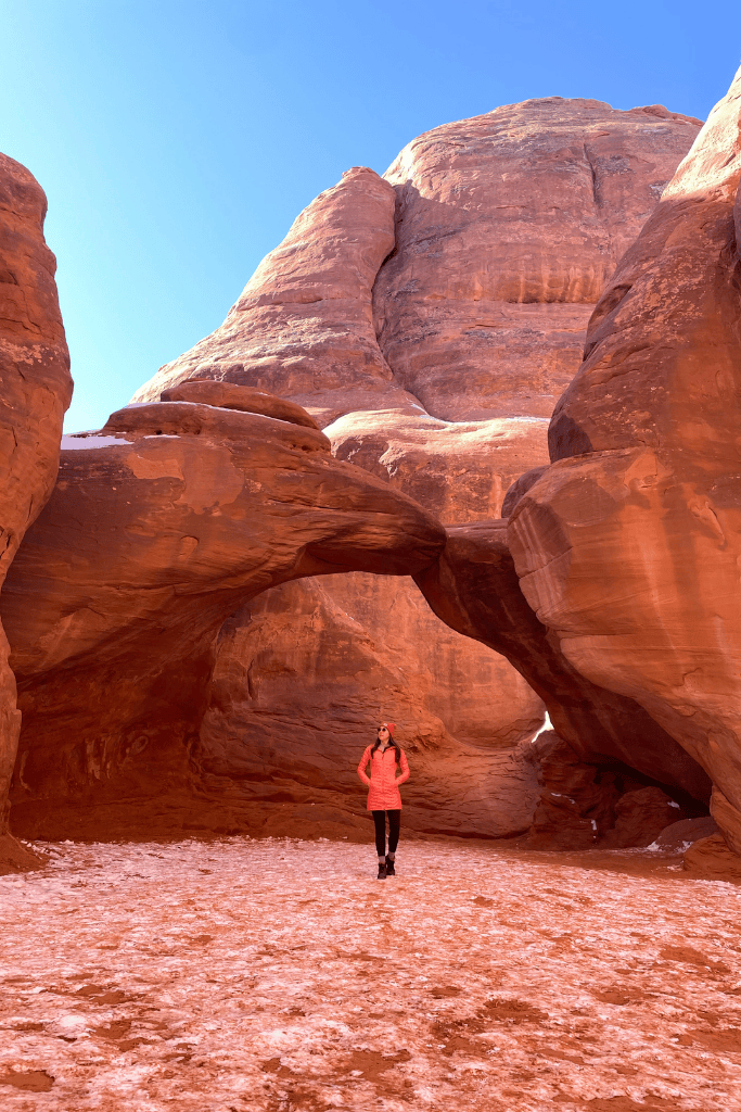 Standing on snow-covered ground, Kate looks up at a natural arch nestled between massive rock formations in Arches National Park.