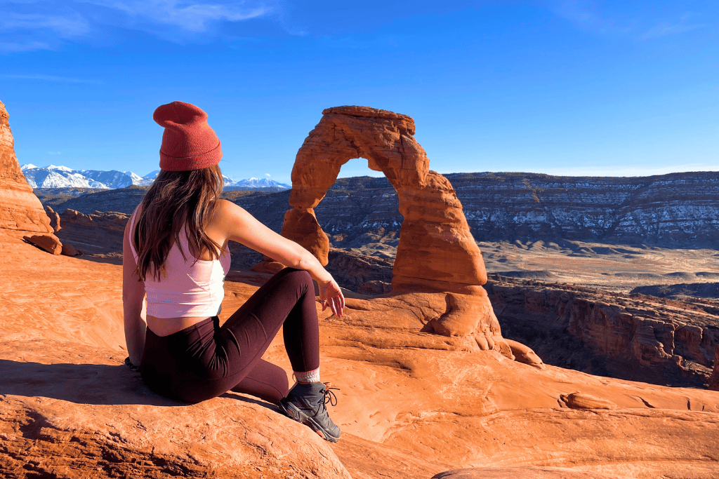 Kate sits on a sunlit red rock ledge, gazing at Delicate Arch with snow-capped mountains and layered desert cliffs in the background at Arches National Park.