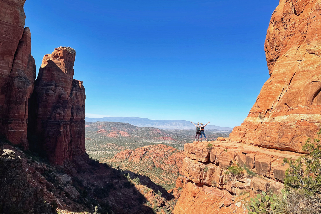 Kate and her friend standing with arms in air on the cliff ledge at the top of Cathedral Rock.