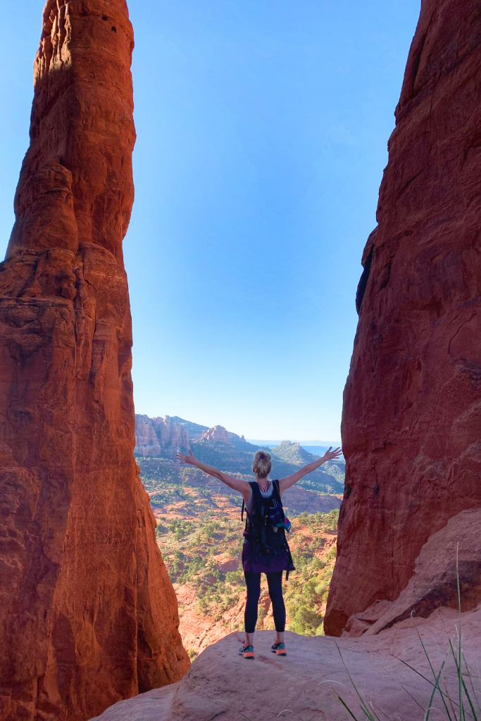 Kate standing with arms reaching to sky with a red rock spire standing tall in front of her. 