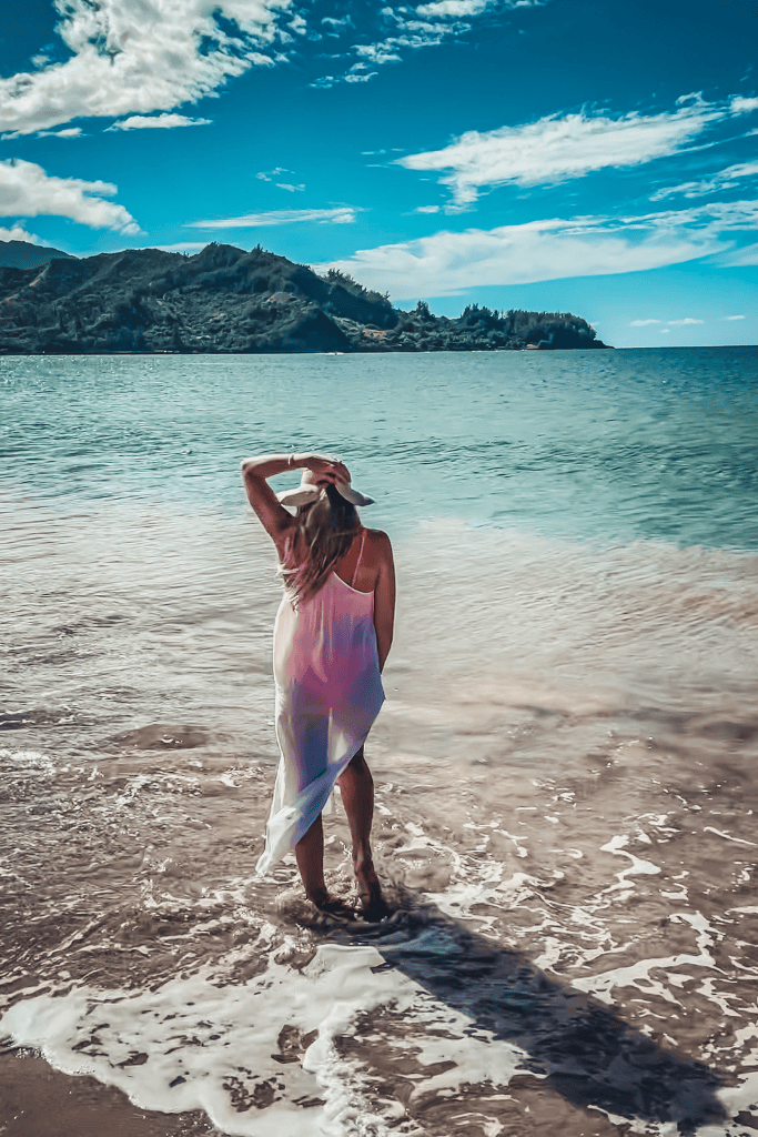 Kate walks into the shallow waves in a sheer white cover-up and sunhat, gazing out at the ocean with Kauai’s lush coastline of the North Shore in the distance.