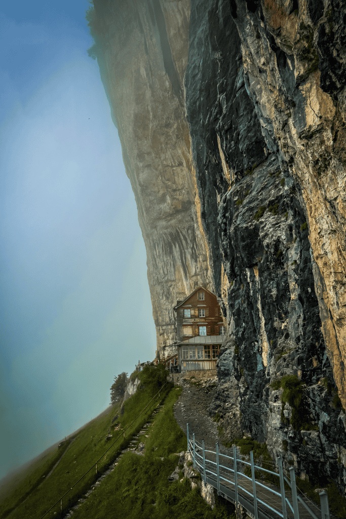 A traditional Swiss mountain house sits tucked beneath a dramatic cliff with a narrow walking path carved into the rock face in Switzerland.