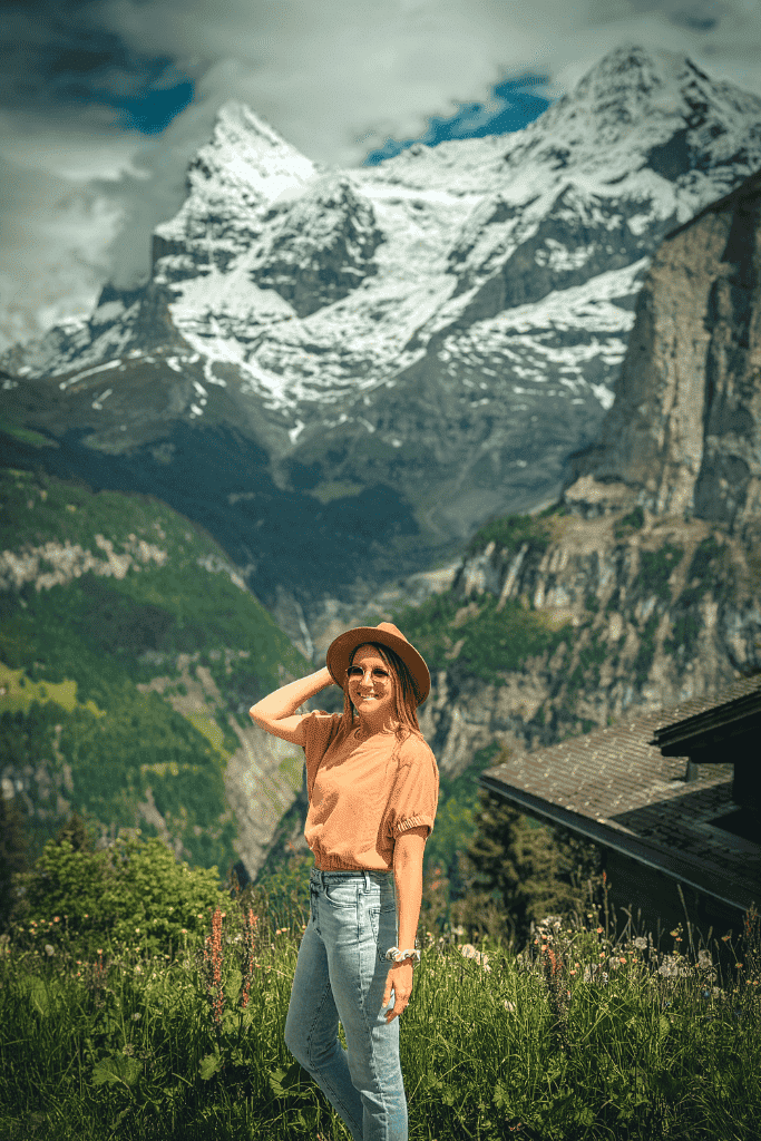 Kate smiles while posing in a wildflower meadow in Mürren with towering, snow-dusted Swiss Alps rising behind her.