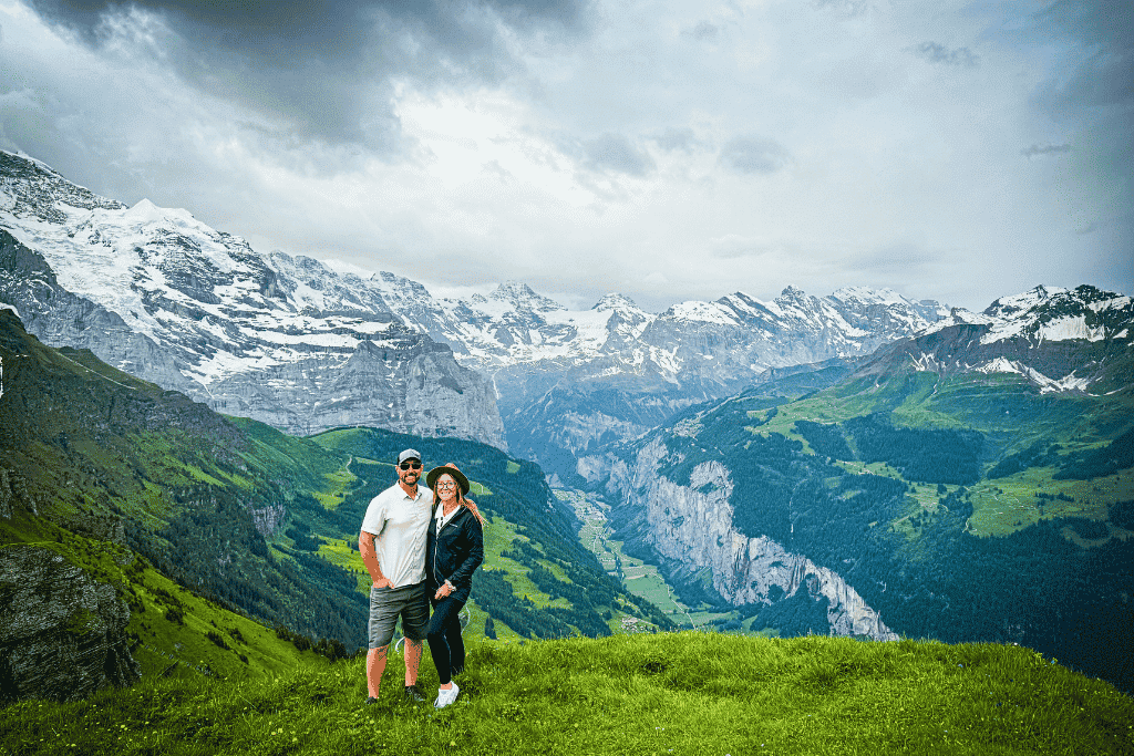 Kate and her husband stand arm in arm on a grassy ridge at Männlichen above Wengen, Switzerland, with sweeping views of the Lauterbrunnen Valley and the snow-capped Bernese Alps behind them.