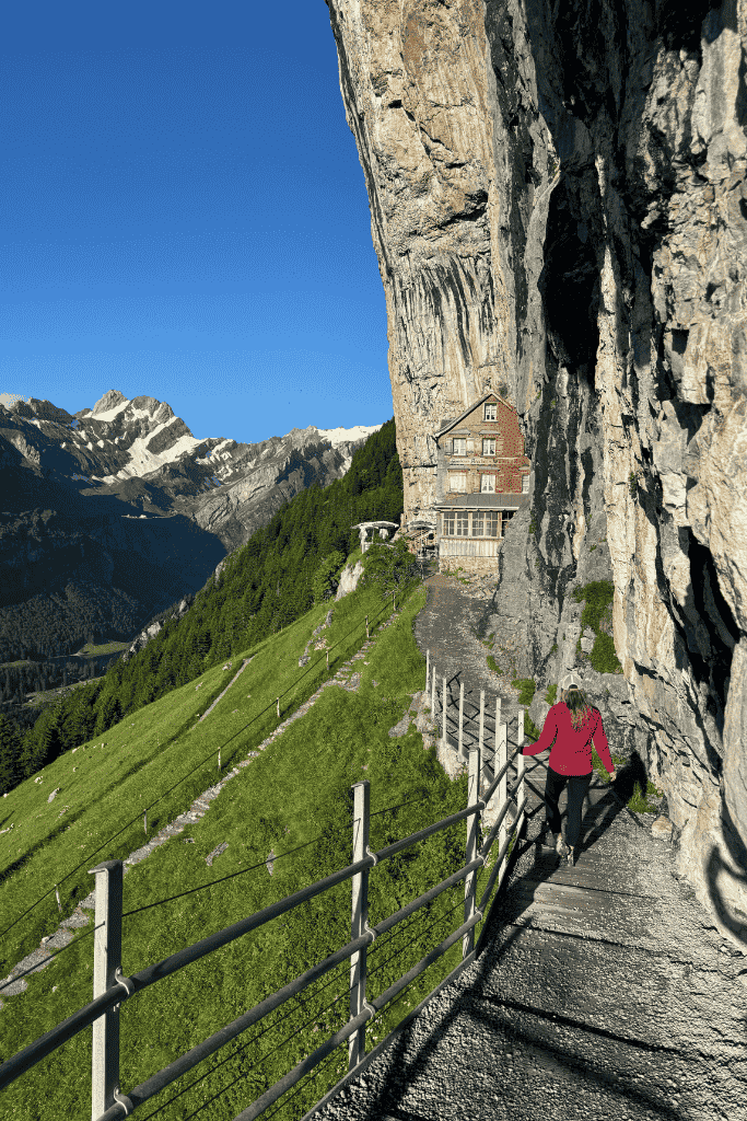 Kate walks down a stone stairway carved into the cliffside toward the Aescher Guesthouse, backed by stunning alpine peaks and clear blue skies.