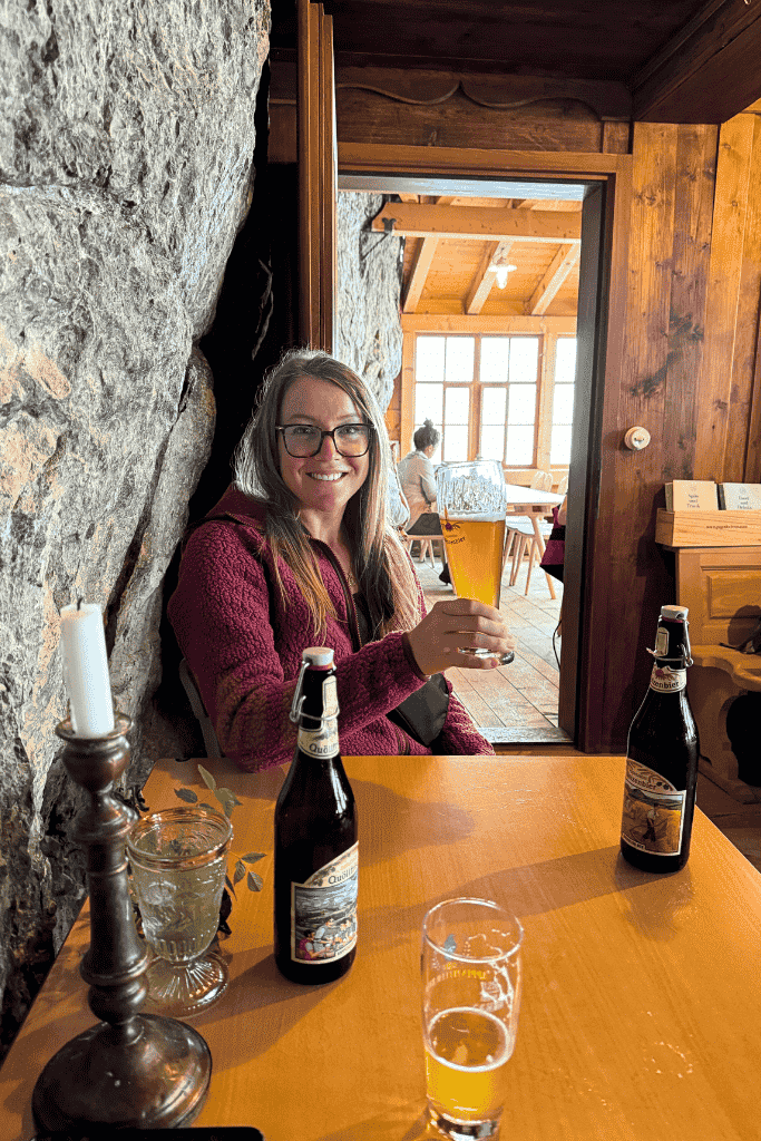 Kate smiles and raises a glass of beer inside the cozy Aescher Guesthouse, seated next to a rock wall and wooden window.