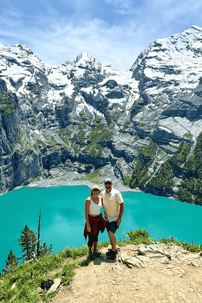 Kate and her husband smile atop a cliff overlooking the vivid turquoise waters of Oeschinensee, framed by towering snow-capped Swiss Alps.