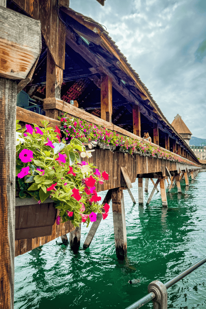 Close-up of Lucerne’s Chapel Bridge adorned with colorful flower boxes, stretching over the turquoise river under a cloudy sky.