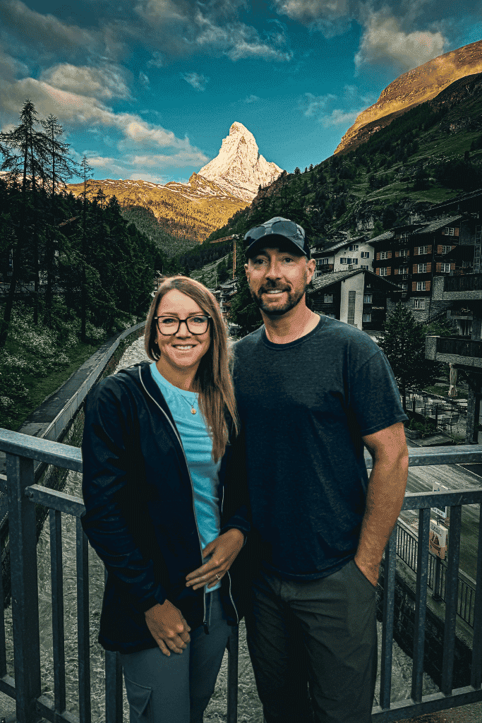Kate and her husband stand on a bridge in Zermatt at sunrise, with the Matterhorn glowing in the background under a vibrant sky.