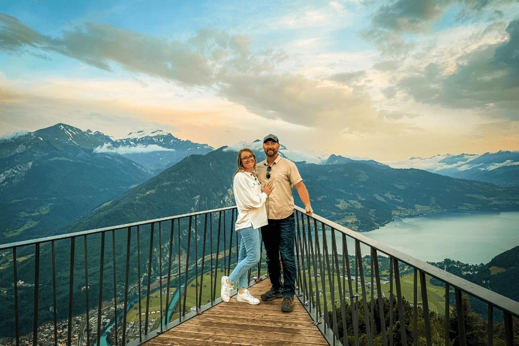 Kate and her husband pose together on a scenic platform high above Interlaken, with panoramic views of alpine peaks, a winding river, and Lake Thun at sunset.