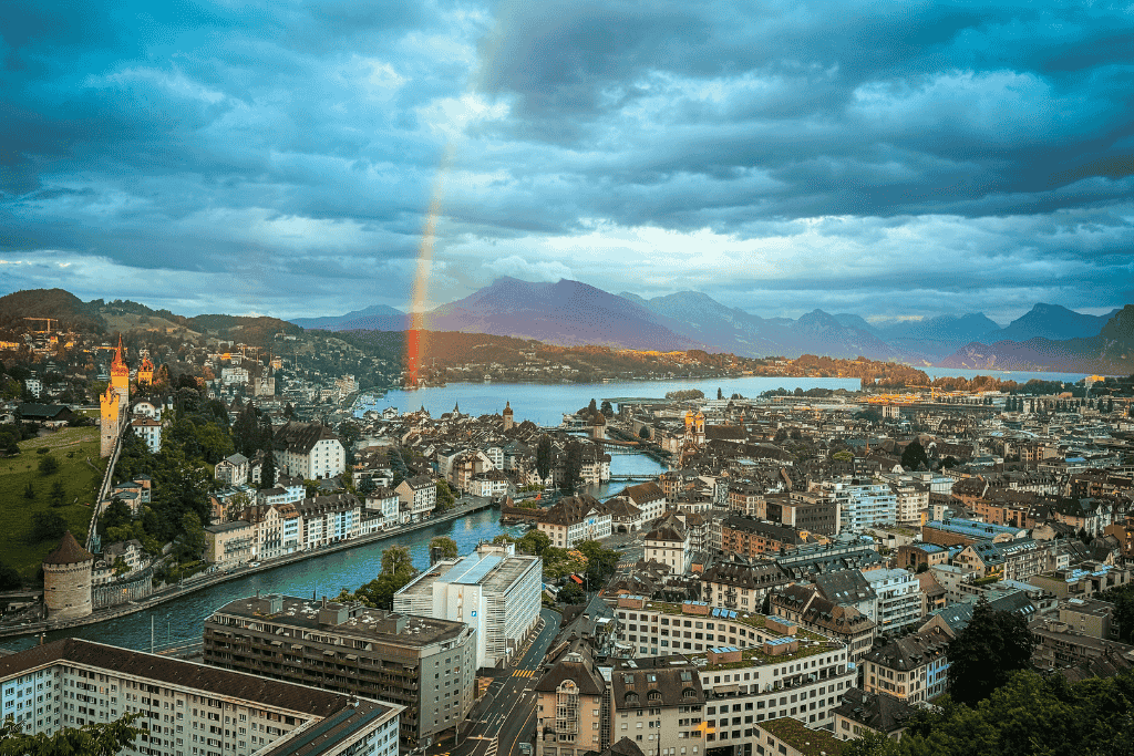 A vibrant rainbow arcs over Lake Lucerne and the old town, illuminating the historic cityscape and mountain backdrop under dramatic evening clouds.