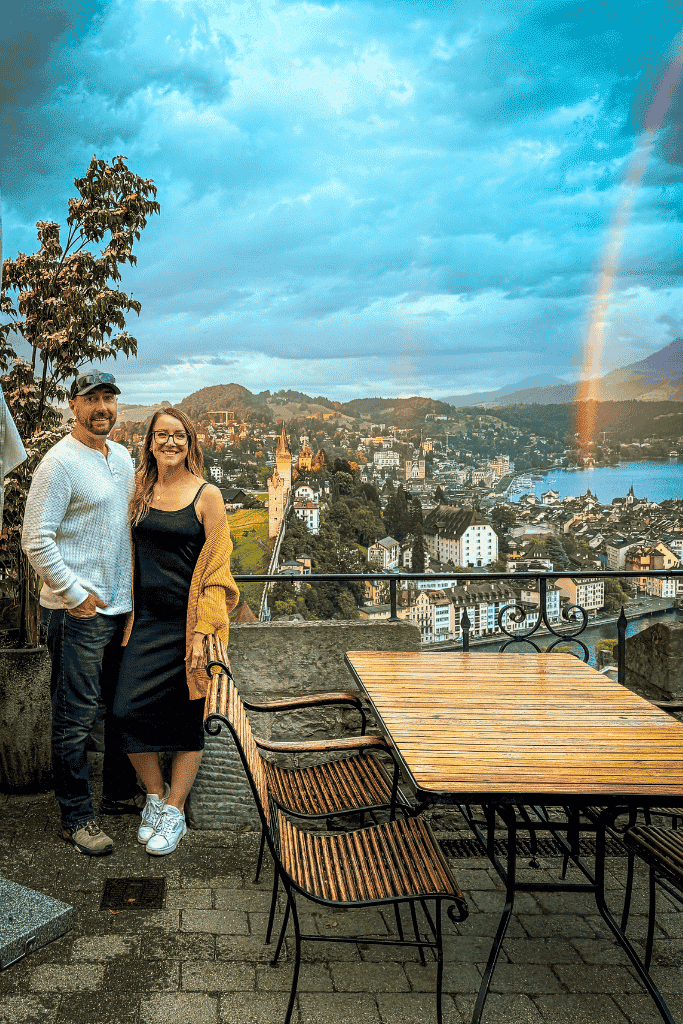 Kate and her husband smile on a terrace overlooking Lucerne’s old town and lake, with a vibrant rainbow arcing across the sky.