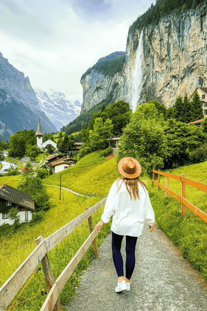 Kate walks along a peaceful path in Lauterbrunnen, Switzerland, with Staubbach Falls cascading down a towering cliff ahead and snow-capped mountains in the distance.