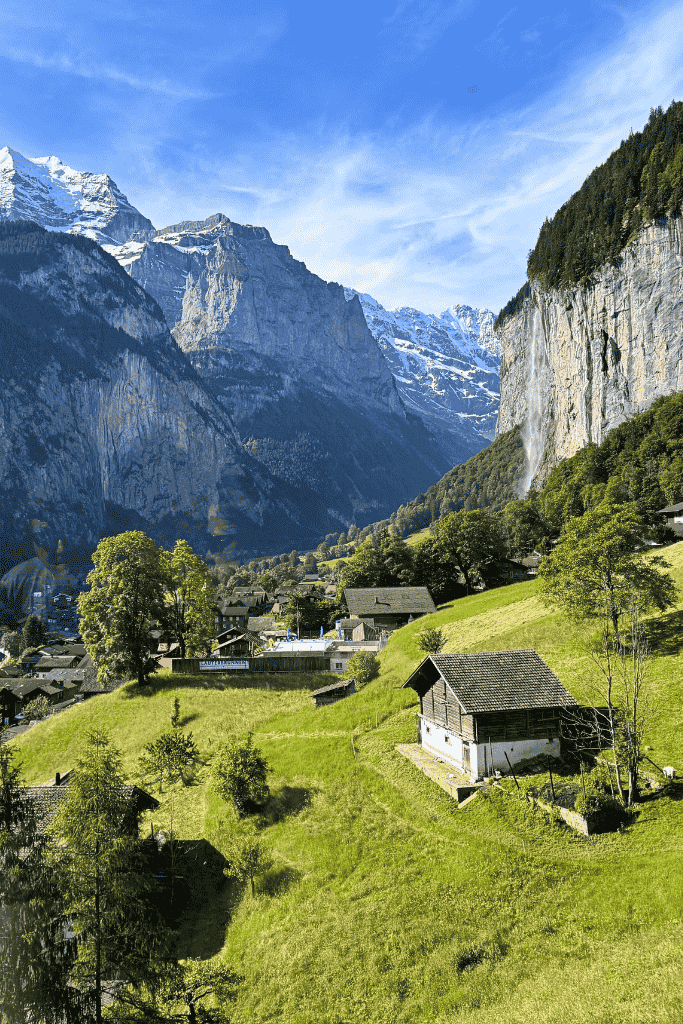 Scenic view of Lauterbrunnen Valley featuring alpine homes, rolling green hills, dramatic cliffs, and the Staubbach Falls under a clear blue sky.