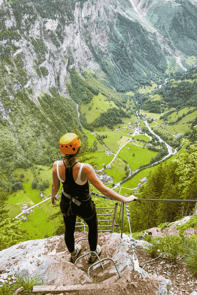 Kate stands at the edge of a metal via ferrata ladder in Mürren, Switzerland, looking out over the steep green Lauterbrunnen Valley far below.
