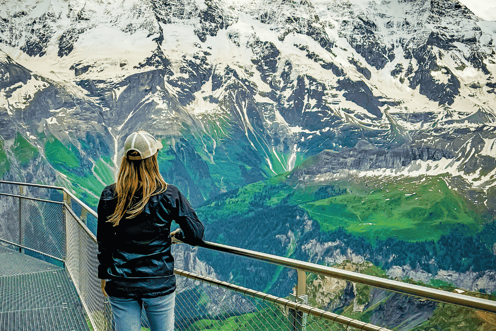Kate is standing on a platform looking out at snow-capped peaks and lush green valleys in Switzerland from a mountain top.