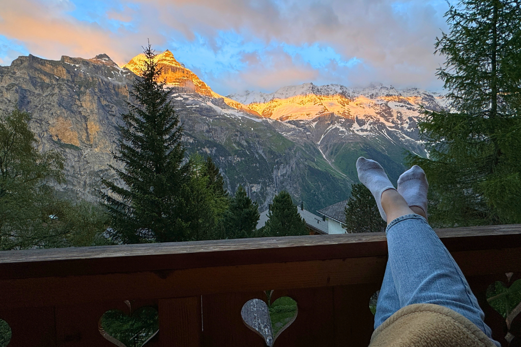 Kate sitting on a balcony with feet propped up looking out at mountains of Switzerland at sunrise.