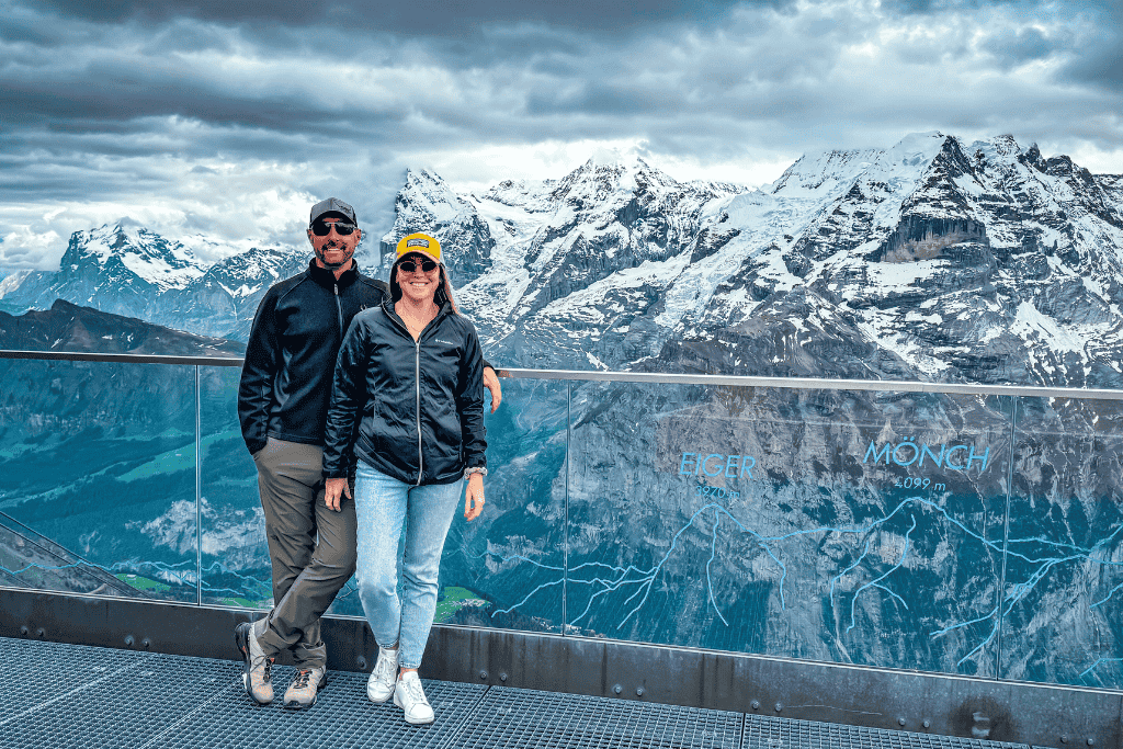 Kate and her husband stand smiling at a scenic Swiss Alps viewpoint with the Eiger and Mönch peaks towering behind them above the valley in Switzerland.