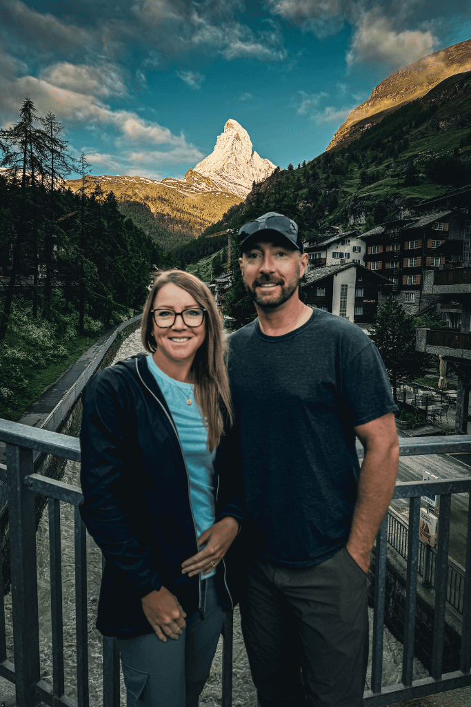 Kate and her husband stand together in a charming Swiss village with the Matterhorn rising dramatically in the background at sunset.