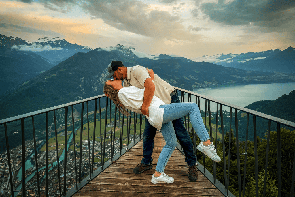 Kate and her husband share a kiss on a scenic overlook above a Swiss town and lake surrounded by rolling mountains at sunset.