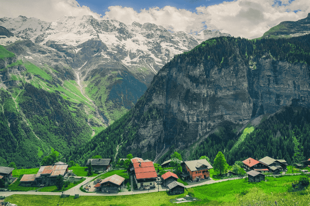 A picturesque Swiss mountain village with traditional chalets sits beneath towering cliffs and snow capped peaks in the Swiss Alps.