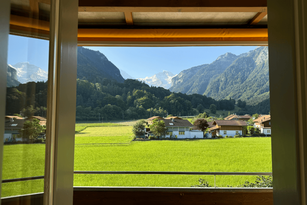 A peaceful view from a Swiss chalet window overlooks bright green fields, traditional homes, and forested mountains in the Swiss countryside.
