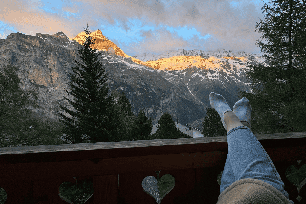 A cozy balcony view in the Swiss Alps shows sock covered feet resting against a railing as golden sunset light hits the snow capped mountain peaks.