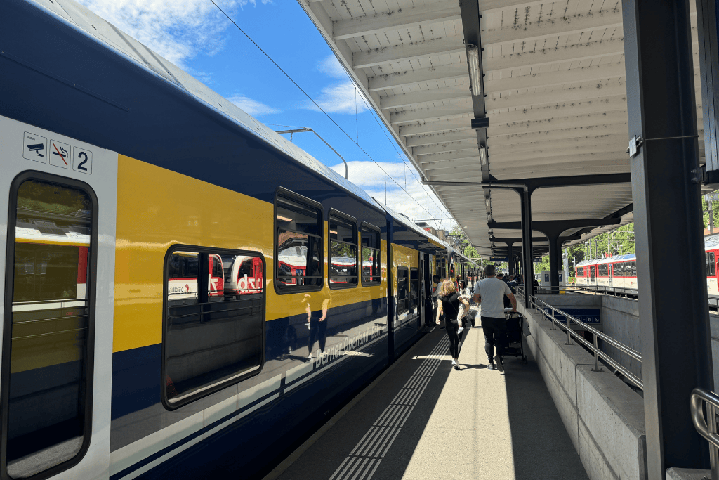 A blue and yellow Swiss regional train waits at a sunny platform as travelers board during a budget friendly Switzerland rail journey.