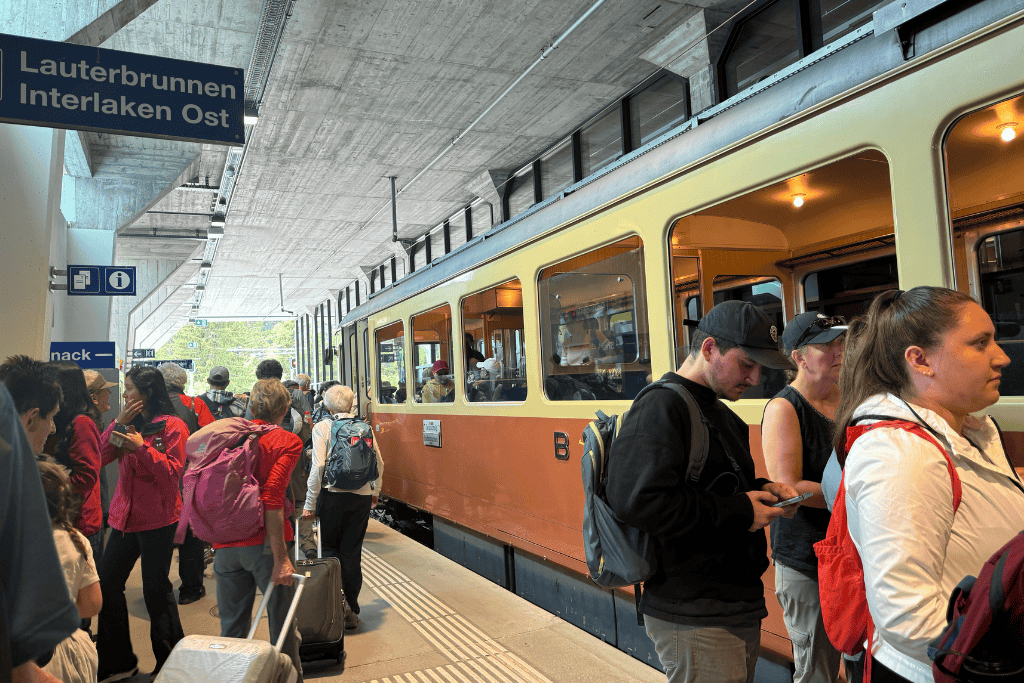 Crowds of travelers with backpacks and suitcases gather beside a train at the Lauterbrunnen and Interlaken Ost station platform in Switzerland.