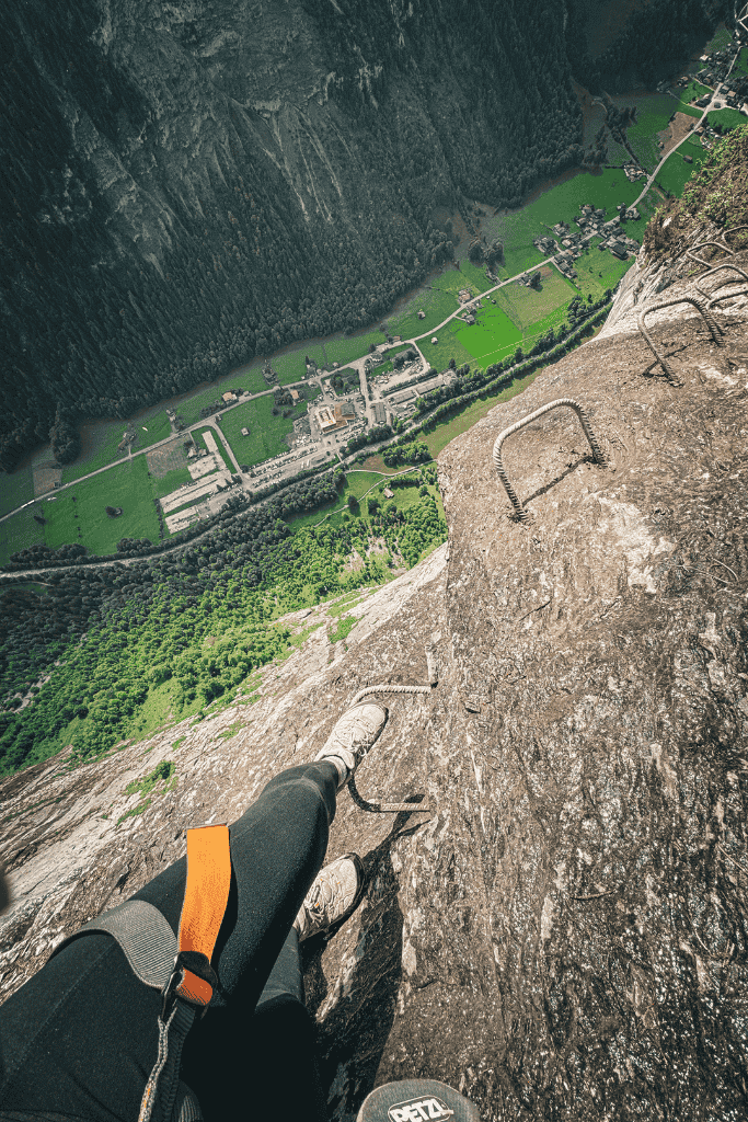 A steep via ferrata route in Switzerland shows metal rungs bolted into a sheer cliff with a dramatic drop to the green valley below.