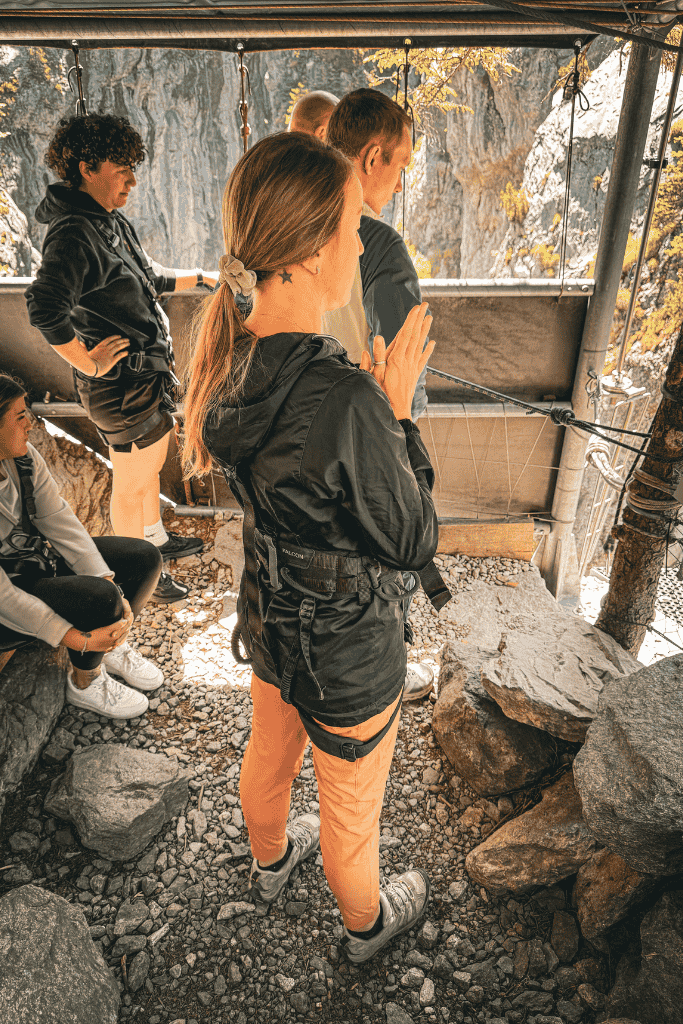 Kate and other climbers pause on a rocky mountain ledge in Switzerland, secured in harnesses as they prepare for the canyon swing.