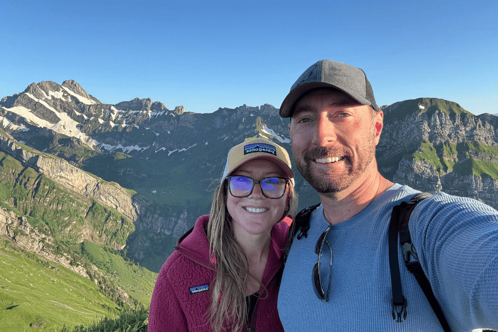 Kate and her husband take a smiling selfie on a mountain overlook with dramatic green cliffs and rugged Swiss Alps peaks behind them in Switzerland.