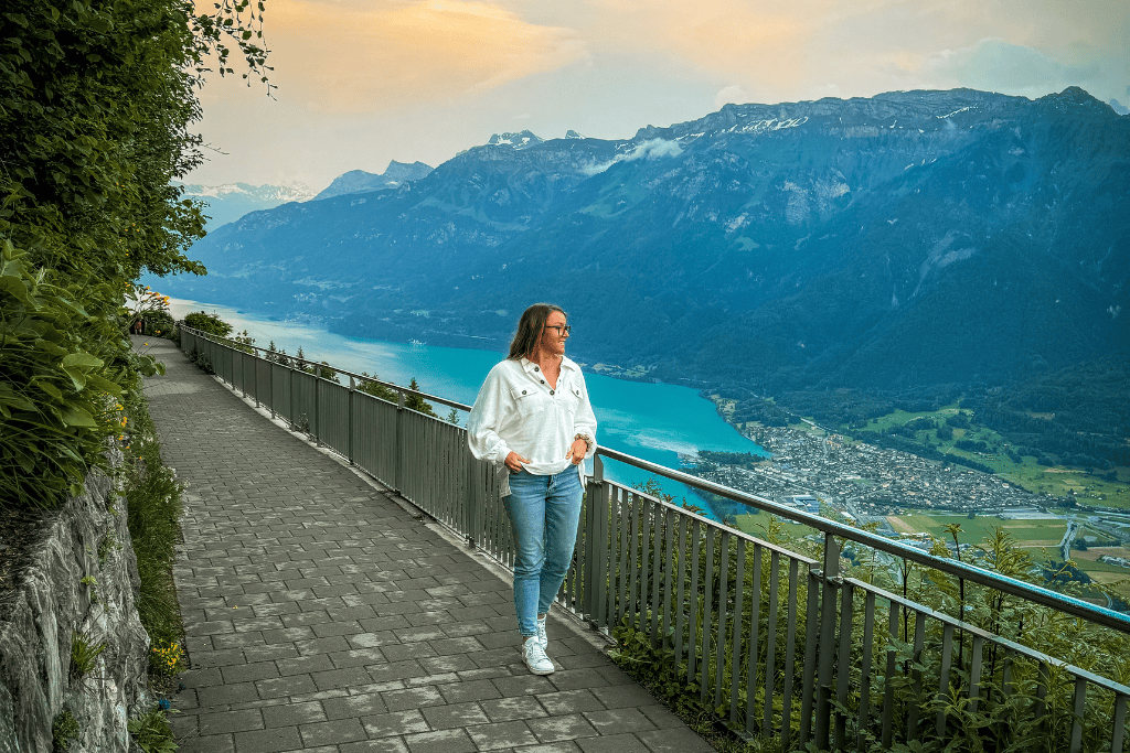 Kate walks along a scenic overlook path above a turquoise Swiss lake with sweeping mountain views at sunset in Switzerland.