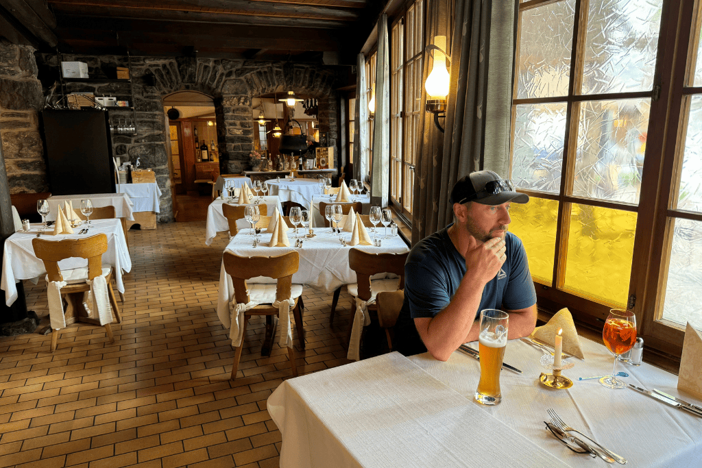 Kate’s husband sits at a cozy Swiss restaurant table with a glass of beer, surrounded by rustic stone walls and neatly set dining tables in Switzerland.