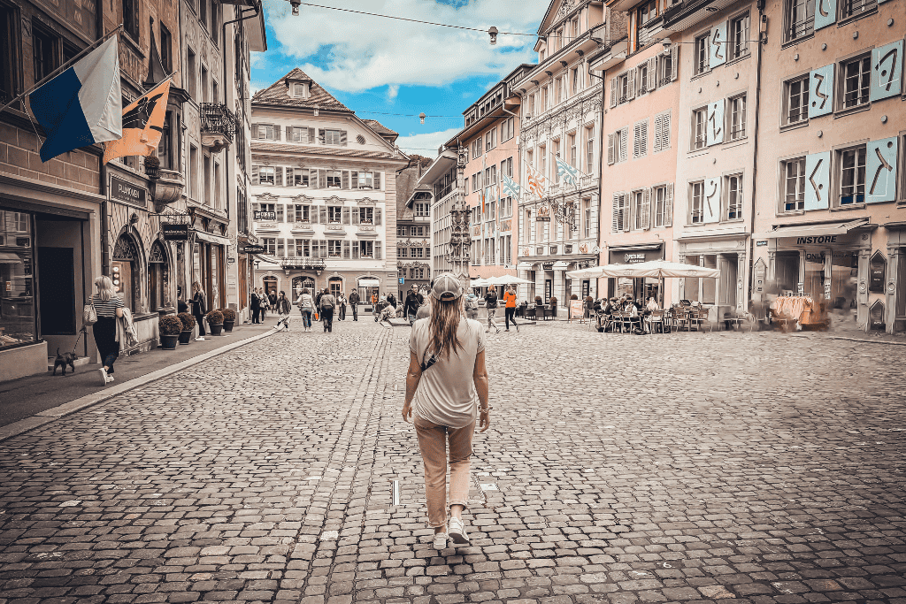 Kate walks through a charming cobblestone old town square lined with historic buildings and outdoor cafés in a picturesque Swiss city.