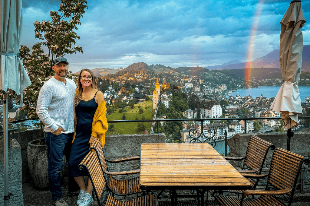 Kate and her husband stand on a scenic terrace overlooking a Swiss lakeside city at sunset with a rainbow stretching across the sky.