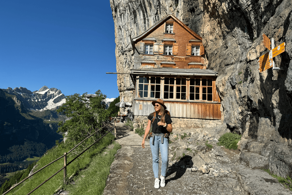 Kate walks along a narrow cliffside path beside a rustic Swiss mountain inn built into the rock face with snow capped peaks rising in the distance.