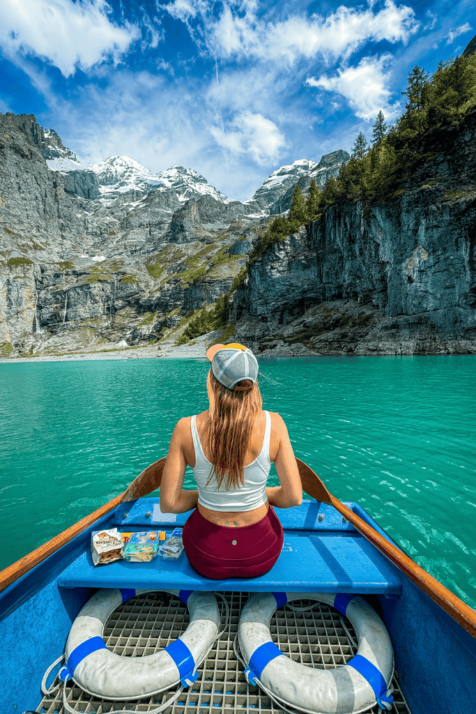 Kate sits at the front of a small boat on a turquoise alpine lake surrounded by towering Swiss Alps peaks and rocky cliffs.