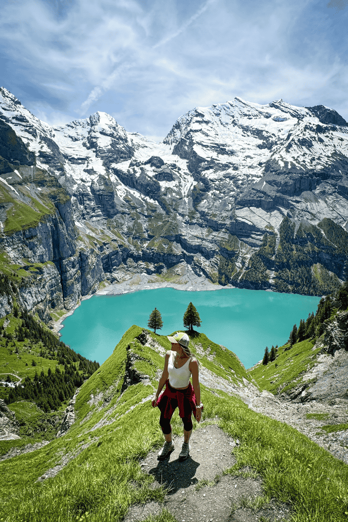 Kate hikes along a narrow ridge trail above a bright blue Swiss mountain lake with dramatic snow capped peaks rising behind her.