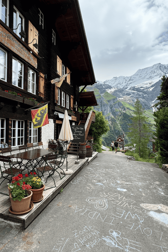 A charming Swiss mountain inn with wooden shutters and outdoor café seating sits along a quiet alpine road beneath towering snow capped peaks.