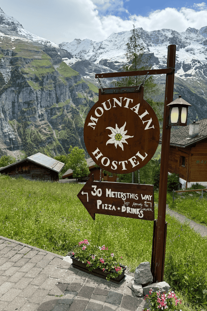 A wooden Mountain Hostel sign with an edelweiss symbol points toward pizza and drinks, set against dramatic Swiss Alps cliffs and green meadows.