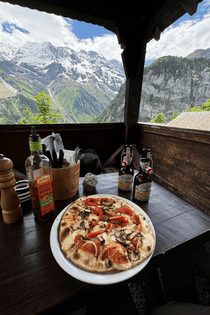 A freshly baked pizza and local Swiss beers sit on a rustic wooden terrace table overlooking a stunning alpine valley and snow capped peaks.