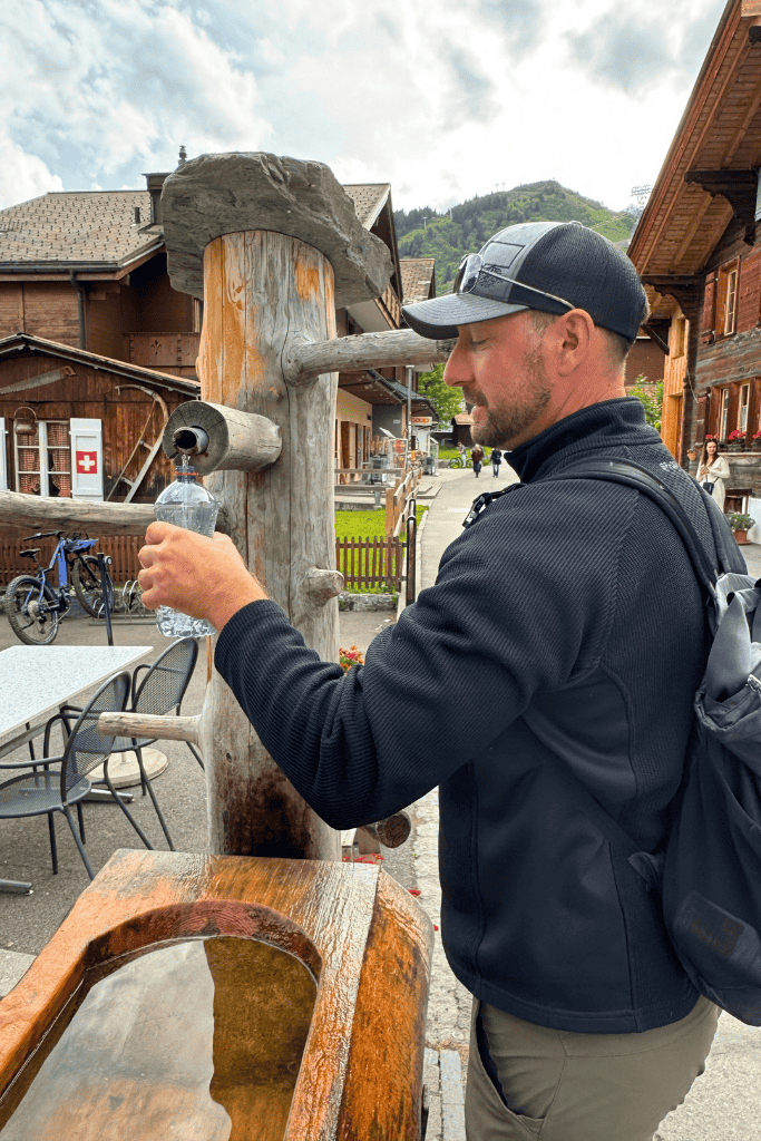 Kate’s husband fills a water bottle at a traditional wooden Swiss village fountain surrounded by rustic alpine buildings.