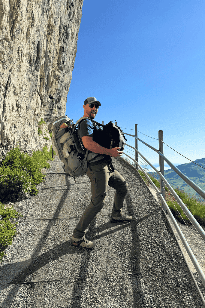 Kate’s husband hikes along a narrow cliffside trail in Switzerland with a heavy backpack and sweeping mountain views in the distance.