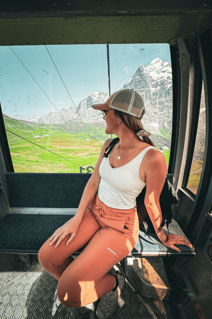 Kate sits inside a mountain cable car in Switzerland looking out at snow capped peaks and green alpine meadows during a scenic Swiss Alps ride.