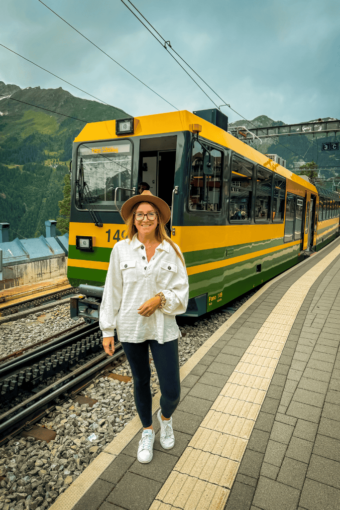 Kate stands on a train platform in Switzerland in front of a bright yellow mountain railway train with alpine peaks in the background.