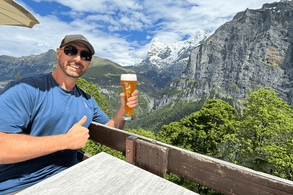 Kate’s husband smiles and holds a glass of Swiss beer on a mountain terrace with dramatic alpine cliffs and snow capped peaks in the background.