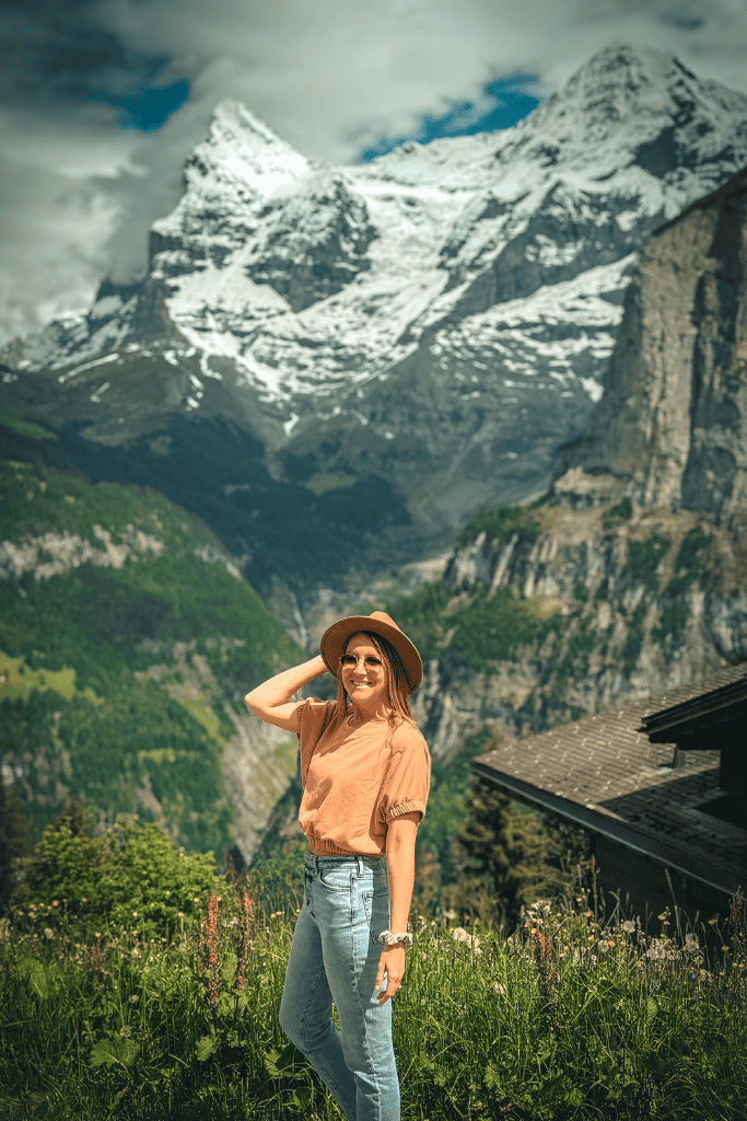 Kate smiles in a wildflower meadow with snow capped Swiss Alps towering behind her during a scenic mountain stop in Switzerland.