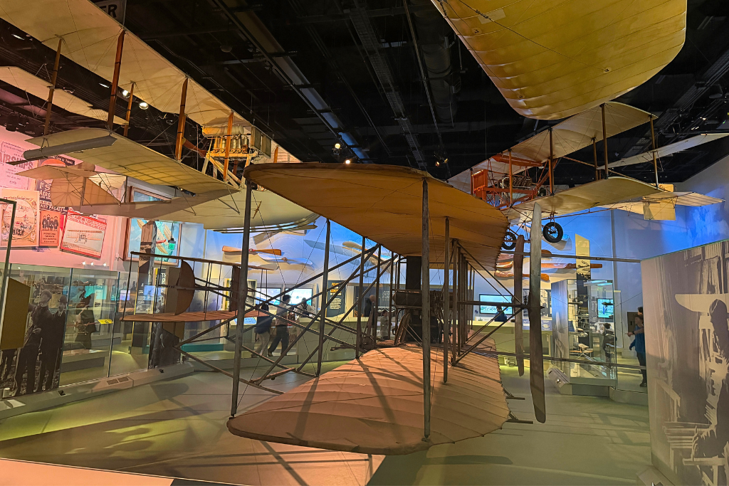 Alt text: Early airplanes with wooden frames and fabric wings hang from the ceiling in a Washington DC museum showcasing the history of flight.